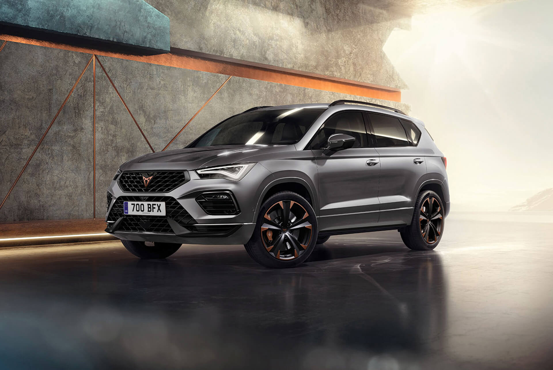 A 2024 cupra ateca in graphite grey, a sporty and compact suv parked in a modern, industrial setting. The car design features aerodynamic lines, led headlights and distinctive alloy wheels with copper accents. The background includes a geometric wall design, contrasting with the sleek vehicle design.