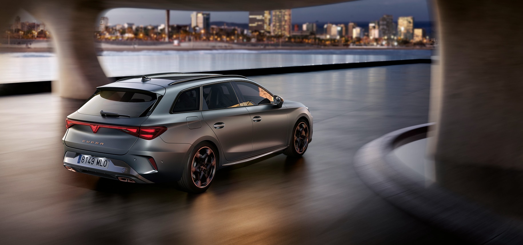 a silver CUPRA Leon Estate driving under a bridge towards a city